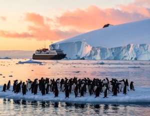 Penguins on an ice floe with a small expedition cruise ship in the background, set against a serene polar landscape at sunrise, showcasing soft pastel skies and towering glaciers.