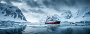 Sleek small expedition ship navigating icy waters surrounded by majestic snow-capped mountains, embodying unique adventure and responsible tourism in remote destinations.